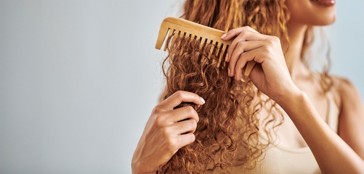 Eine junge Frau mit voluminösen rot-braunen Locken kämmt mit einem sehr groben Holzkamm ihre Haare. Nur die Haare und Hände sind zu sehen.